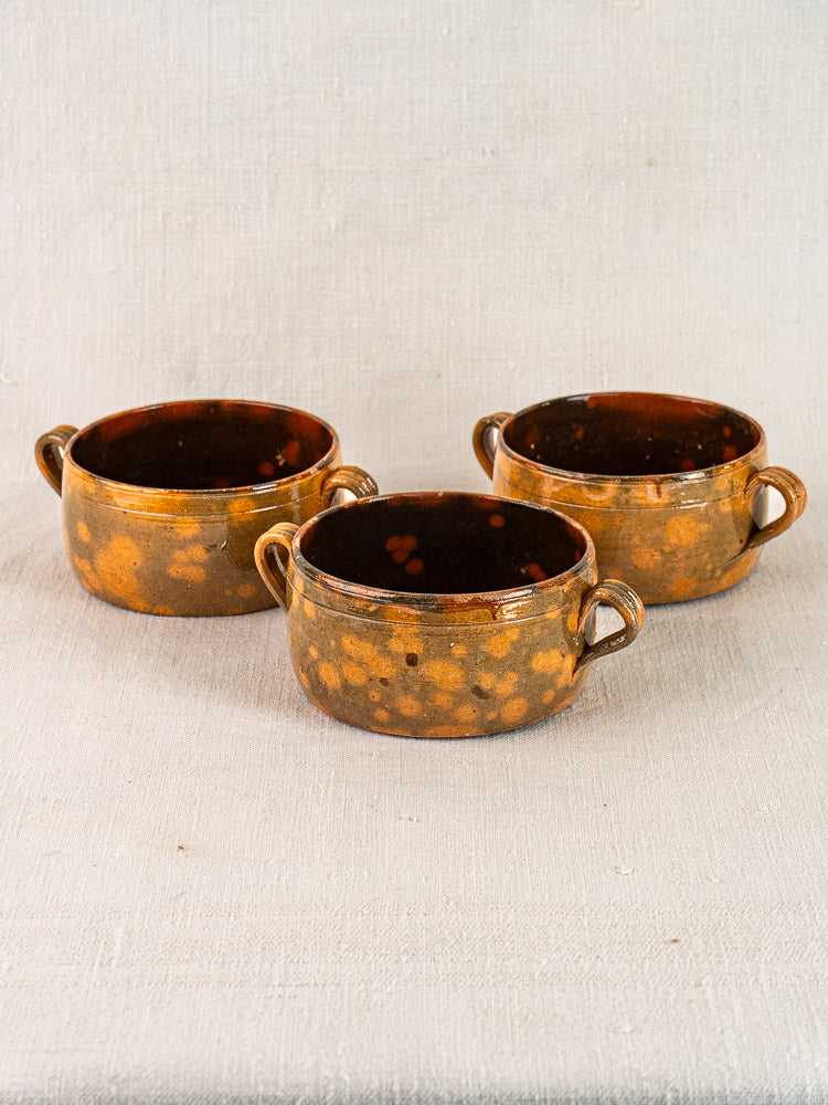 Set of three vintage French Vallauris earthenware bowls with double handles, rustic brown and yellow glaze on textured fabric background.