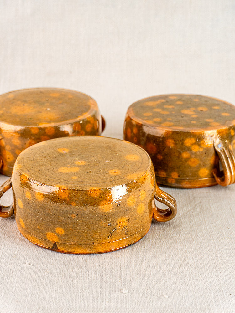 Set of three vintage French Vallauris bowls with orange-brown mottled glazed bases and twin handles, on textured light fabric.