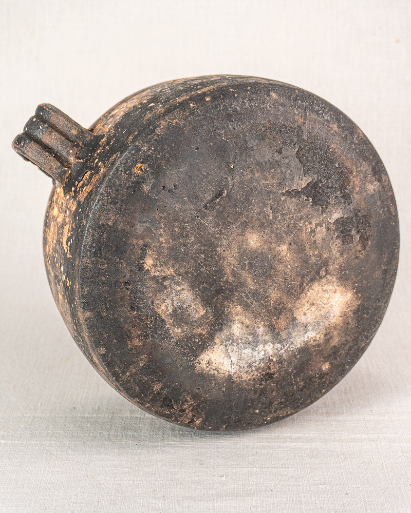 Early 20th-century French Vallauris glazed terracotta pot with three short spouts, worn blackened surface, on light fabric.
