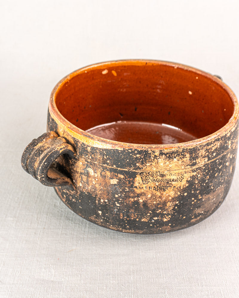 Early 20th century French Vallauris terracotta cooking pot with two handles, weathered exterior, and glossy brown interior on light fabric.