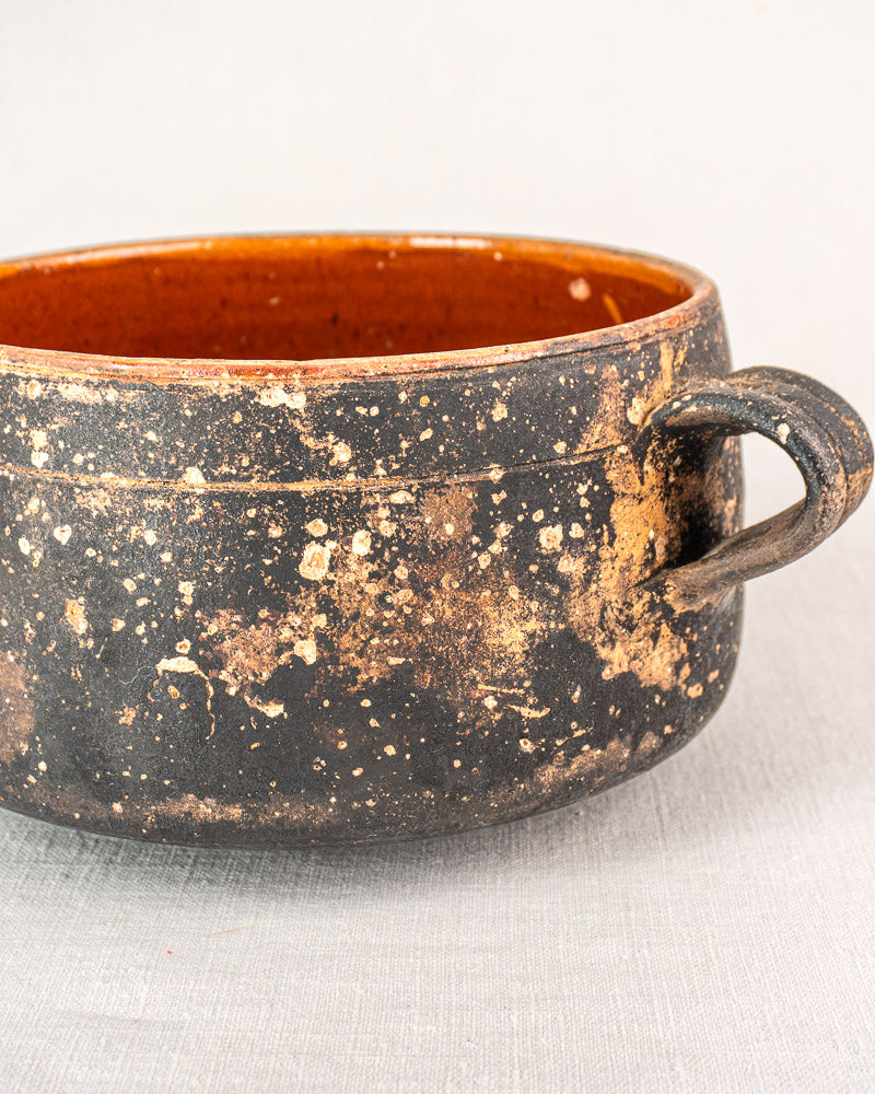 French Vallauris glazed terracotta cooking pot with single handle, speckled black-brown exterior, on light fabric surface.