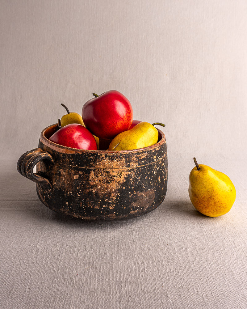 Vintage French Vallauris glazed terracotta cooking pot with red apples and yellow pears on light gray fabric.