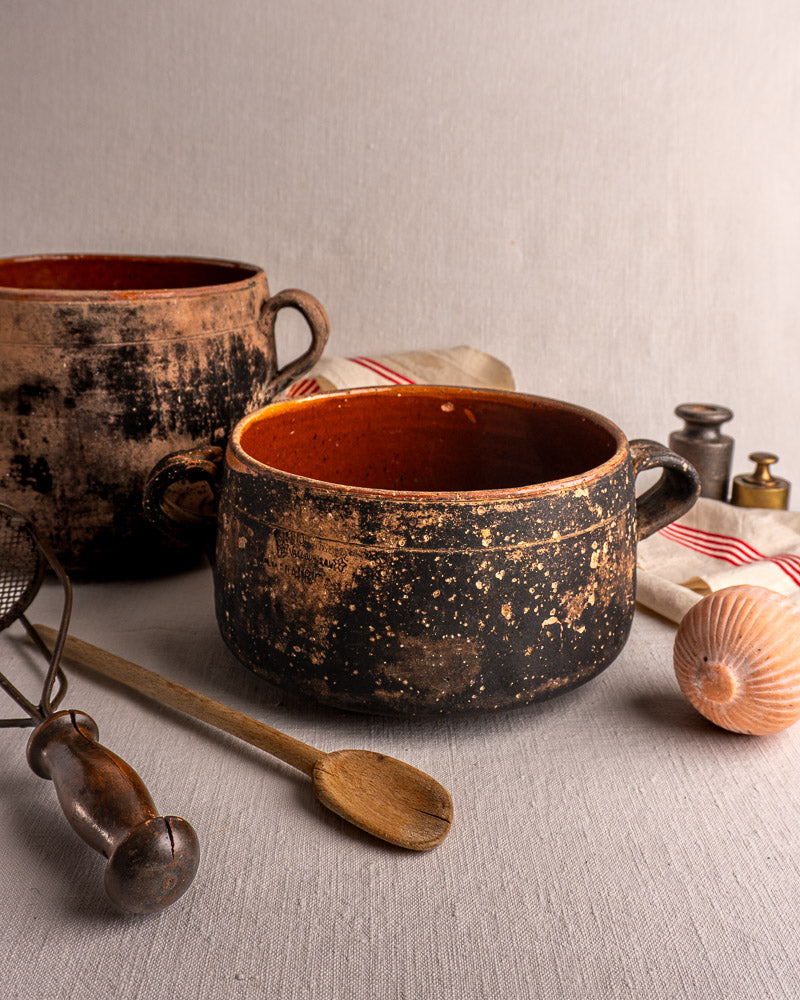 Two early 20th-century French Vallauris glazed terracotta cooking pots with spoon, whisk, and striped cloth on light fabric.