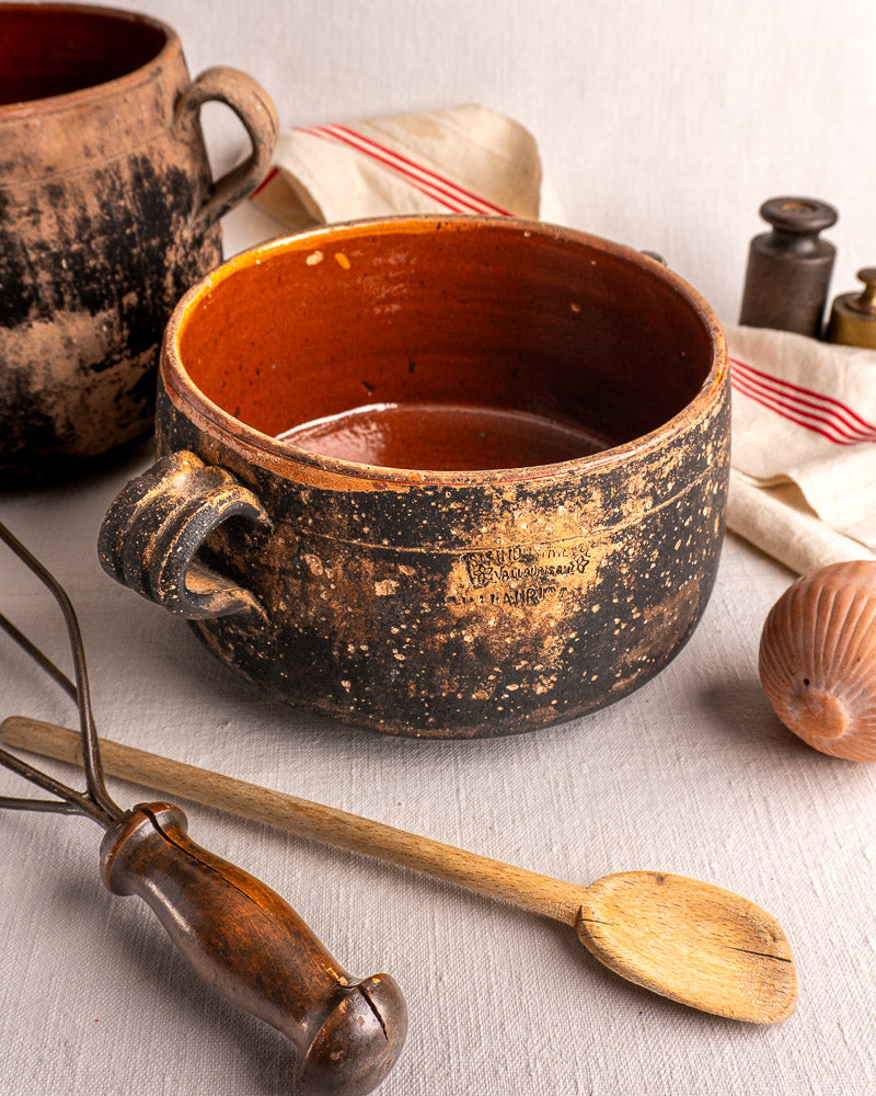 Vintage French Vallauris glazed terracotta cooking pot with handle, on cloth beside wooden spoon, whisk, onion, and striped towel.