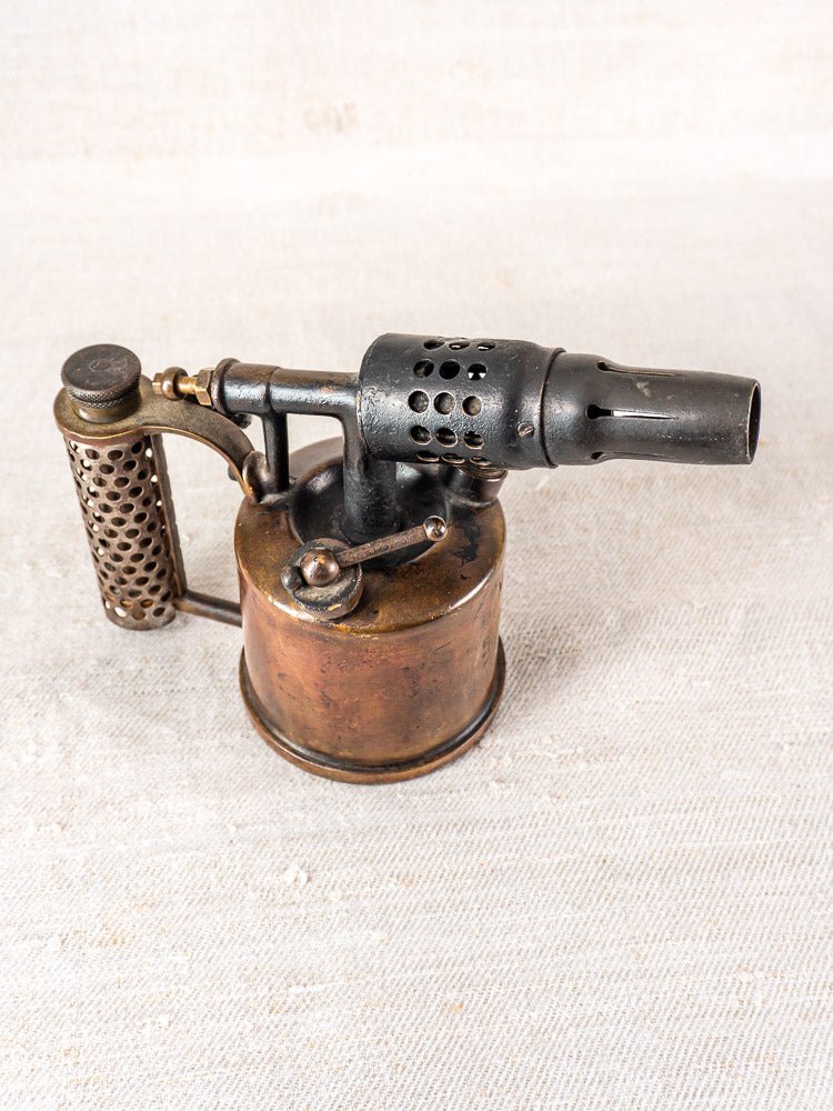 Antique French Vesta F.J. Paris blowtorch, brass tank and metal nozzle, on textured surface.