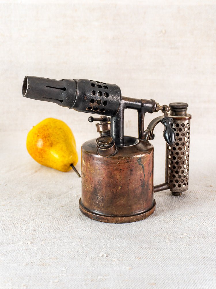 Vintage French Vesta F.J. Paris blowtorch with worn metal body on light fabric, yellow pear behind.