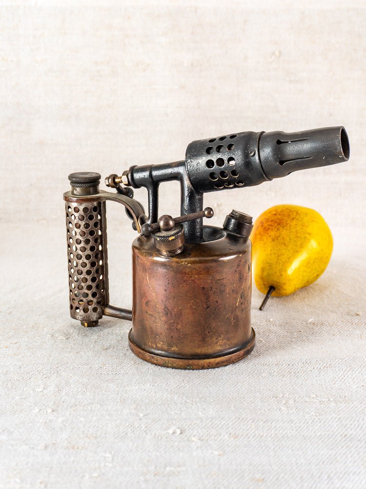 Vintage French Vesta F.J. Paris blowtorch with perforated handle, circa 1920, beside a yellow pear.