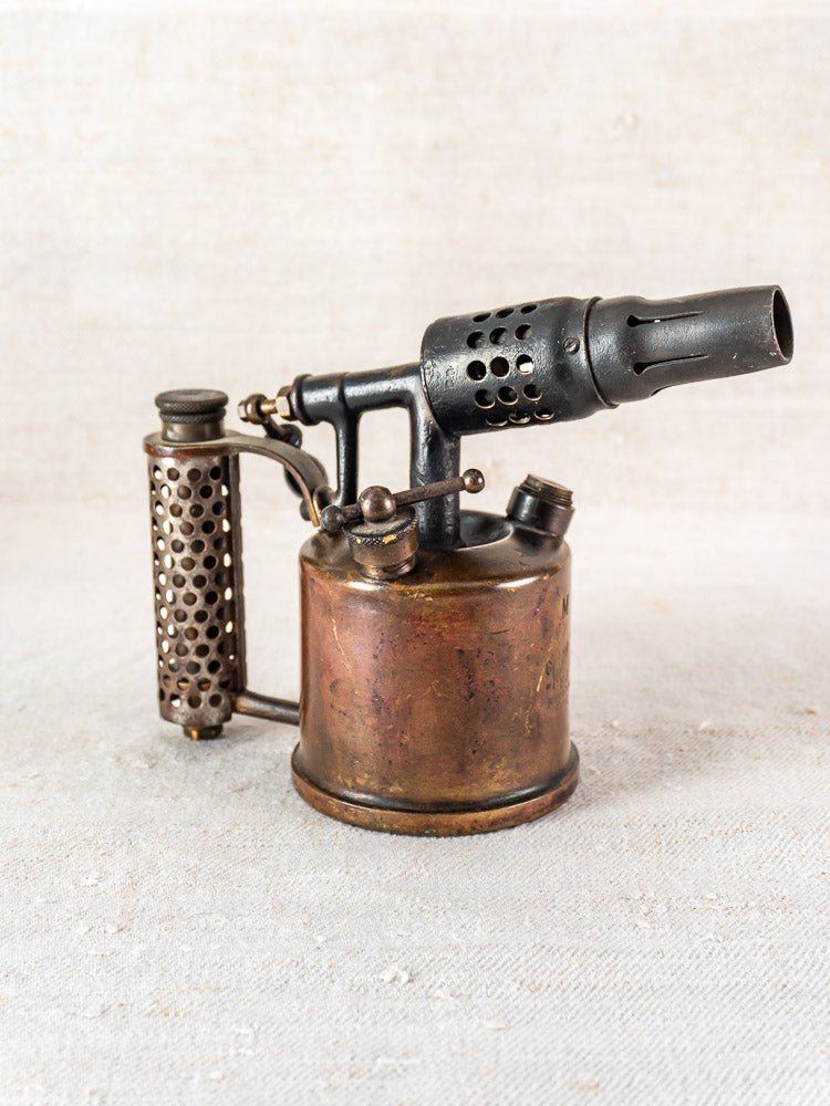 Vintage French blowtorch by Vesta F.J. Paris, circa 1920, with cylindrical handle and perforated casing on light textured surface.
