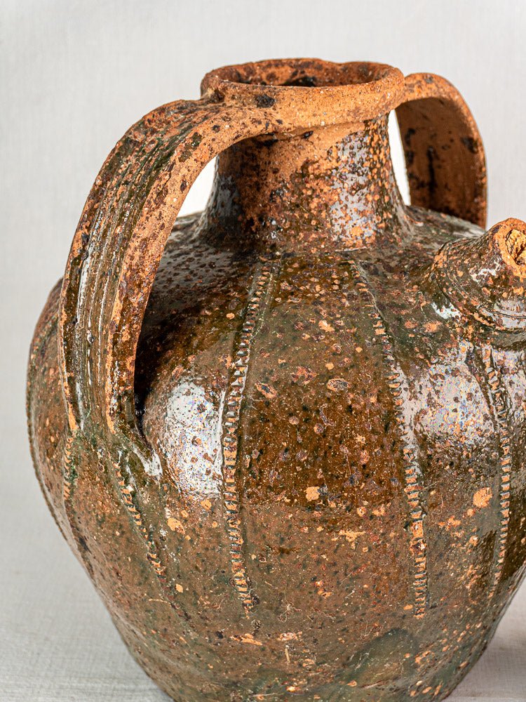 Antique French terracotta walnut oil cruche with green-brown glaze, ribbed design, two handles on neutral fabric.