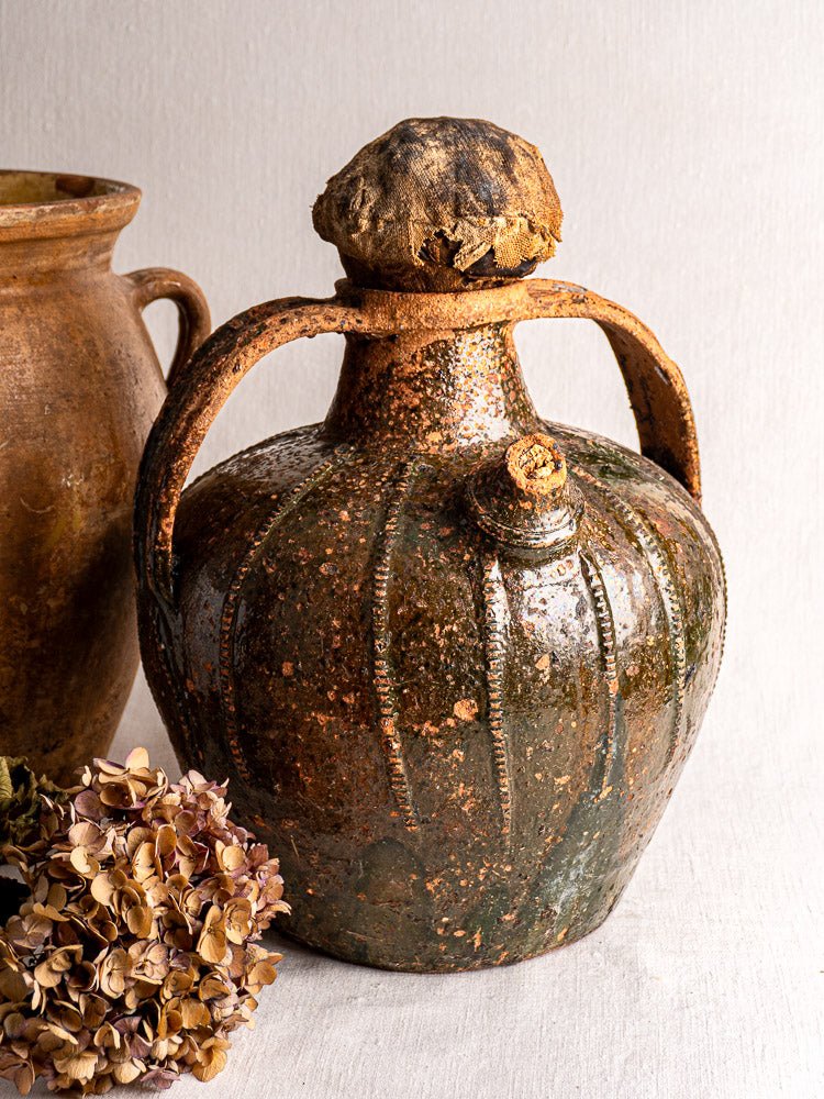 Antique French glazed terracotta cruche with rustic brown finish beside dried hydrangea on fabric.