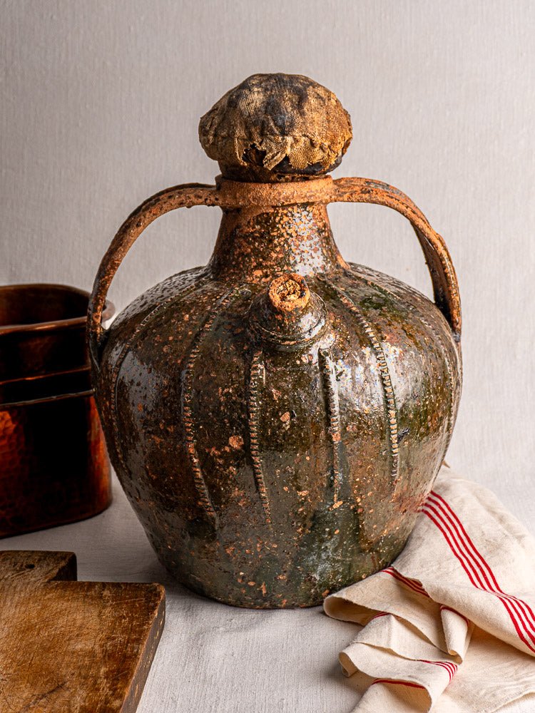Antique French terracotta walnut oil cruche with double handles and wrapped stopper on rustic table.