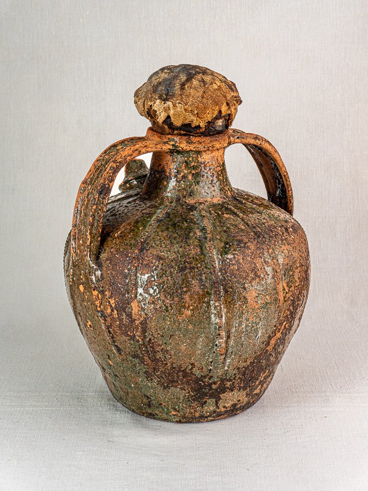 Antique French terracotta oil cruche with two handles, earthy glaze, cork stopper; plain background.