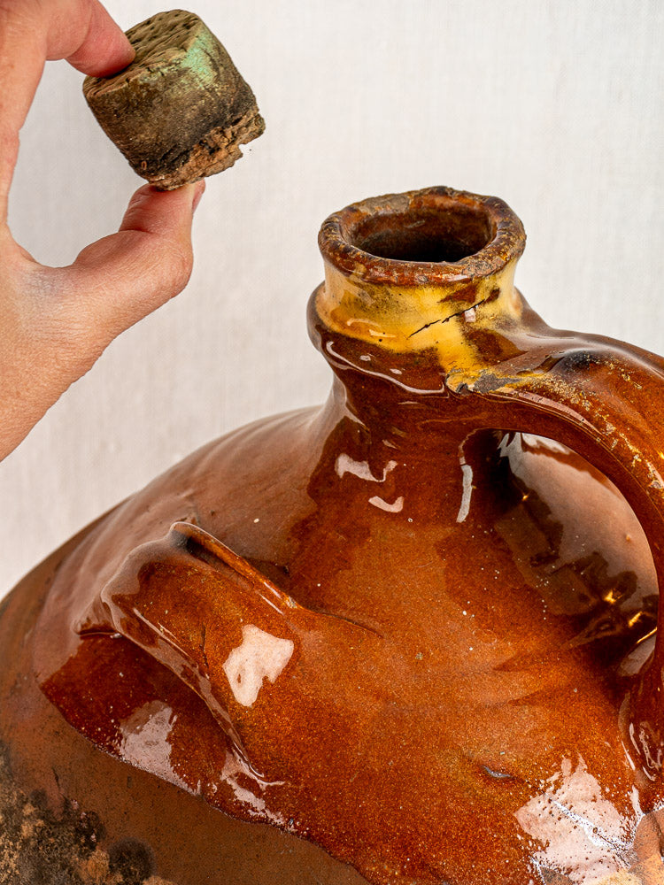 Antique French Auvergne terracotta oil jar with handle and cork stopper on a light background.