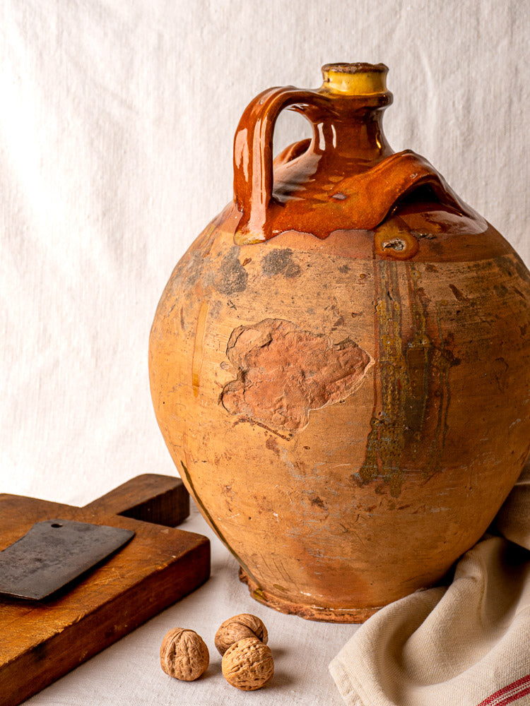 Antique French terracotta walnut oil jar with original cork, shown beside cutting board and walnuts.