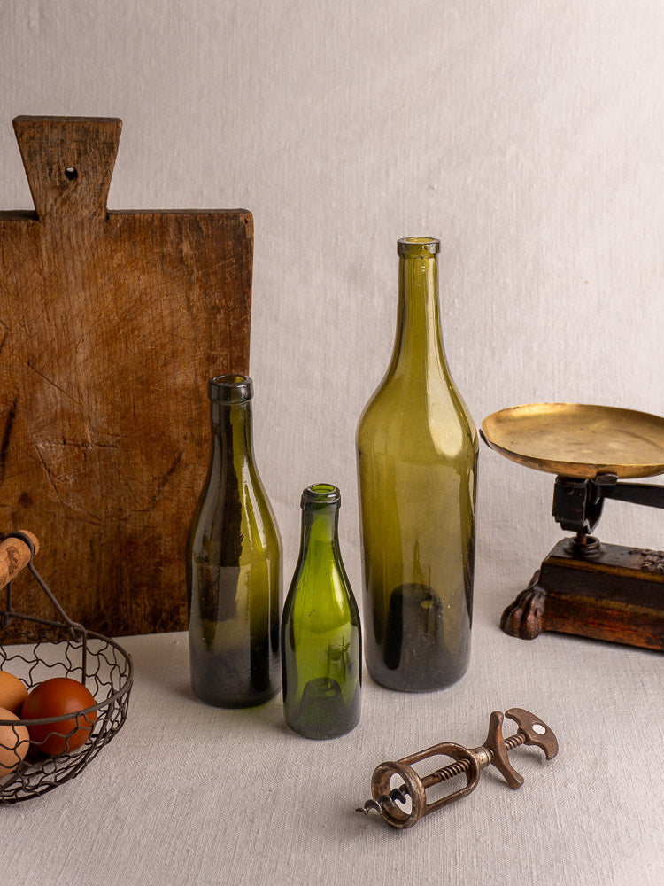Set of three antique French mid-19th century hand blown wine bottles with vintage scale and kitchen props on neutral fabric.