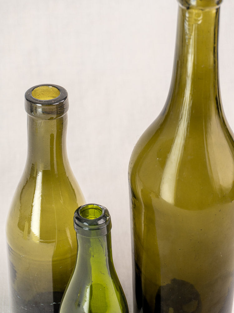 Set of three mid-19th century French hand blown olive green glass wine bottles in assorted sizes on a light background.
