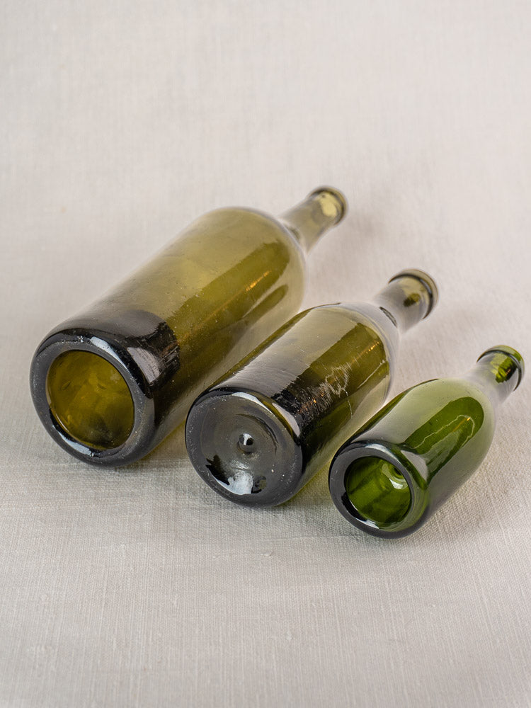 Set of three mid-19th century French hand blown olive green glass wine bottles on light textured surface.