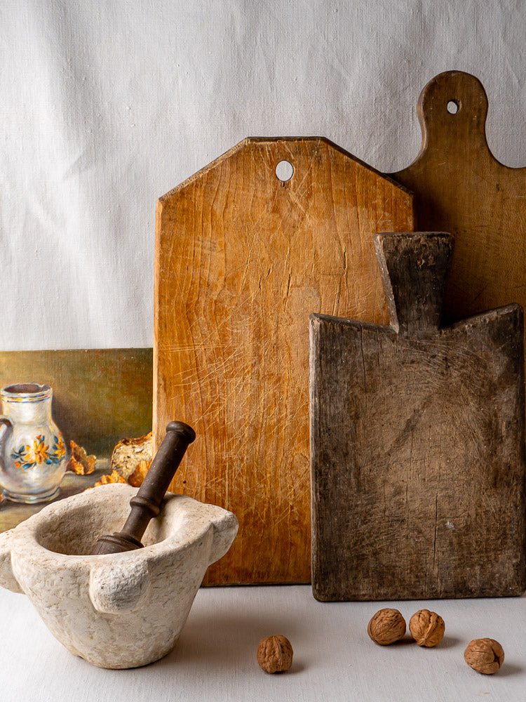 Antique French wooden cutting boards with stone mortar and pestle, walnuts, and floral jug painting.