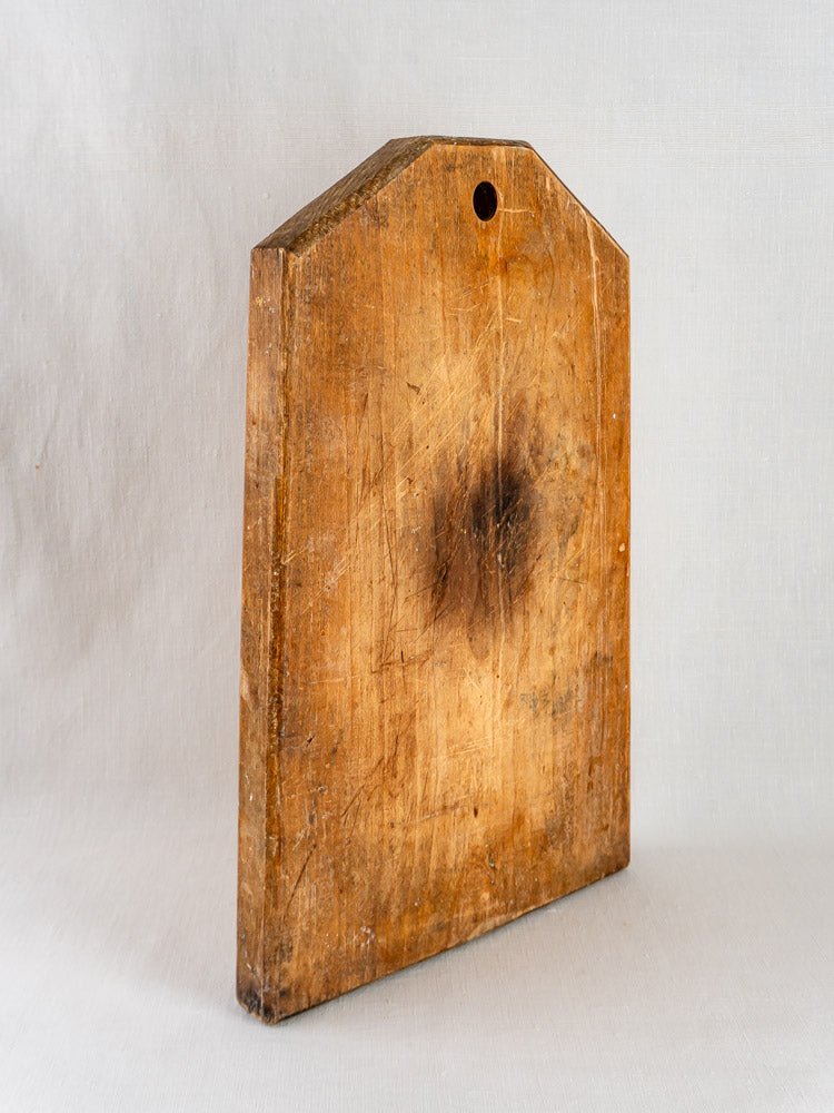 Antique 19th-century French wooden cutting board with knife marks, dark central stain, and hanging hole.