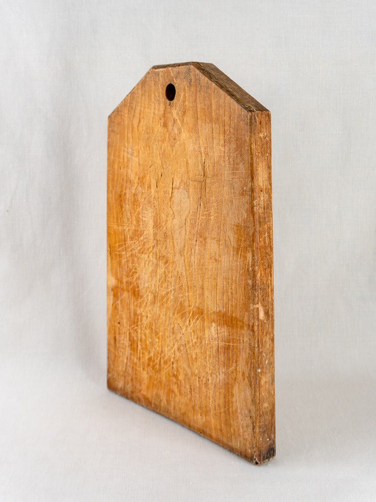 Antique 19th-century French wooden cutting board with beveled top edge and hanging hole, upright against a light background.