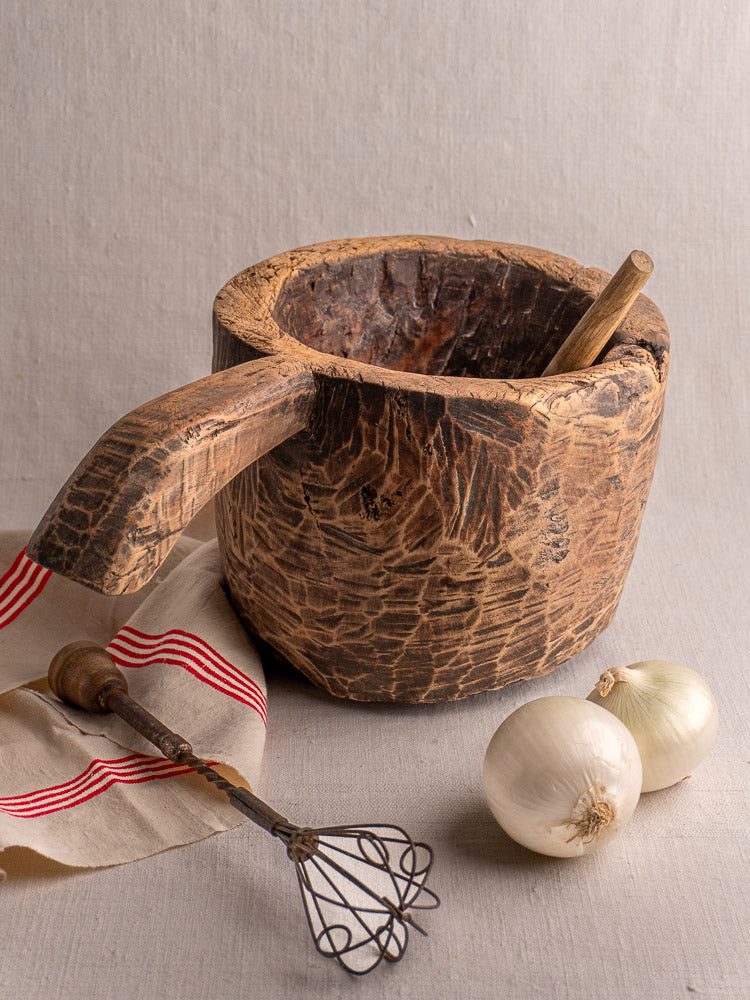 Antique French wooden mortar and pestle, hand-carved, shown with whisk, cloth, and onions.