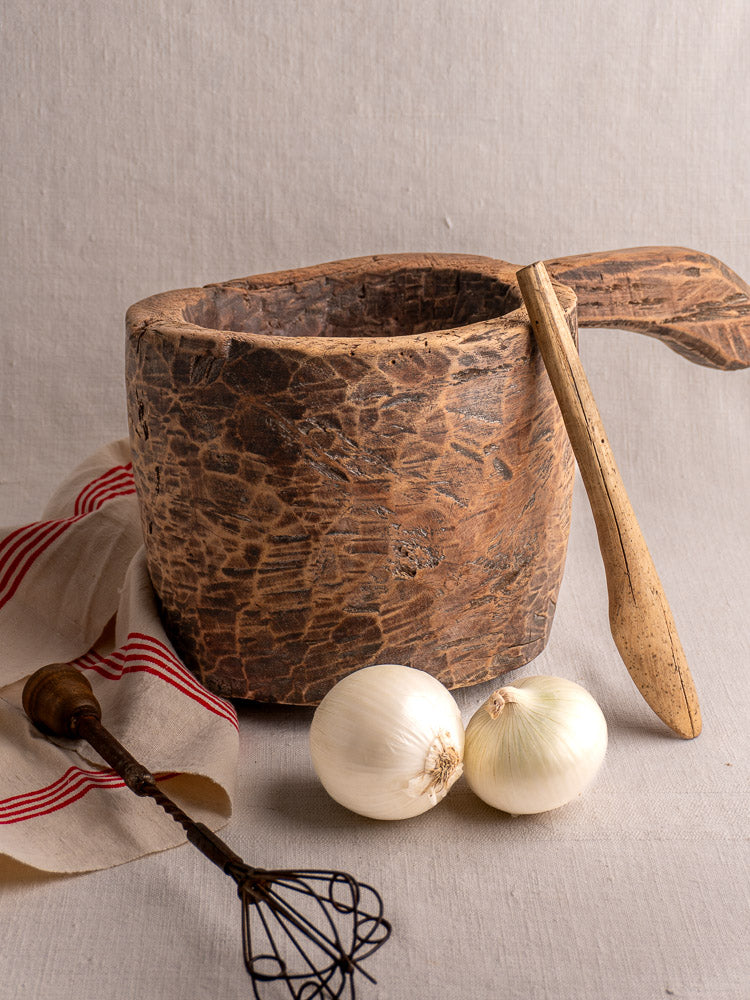 Antique French wooden mortar and pestle on light cloth with onions, whisk, and striped towel nearby.