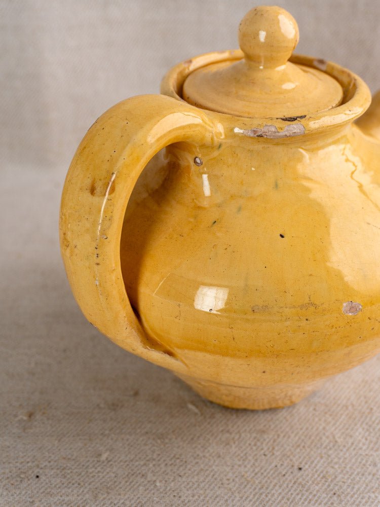 Antique French yellow glazed earthenware teapot with round lid and large handle, minor wear, on beige fabric.