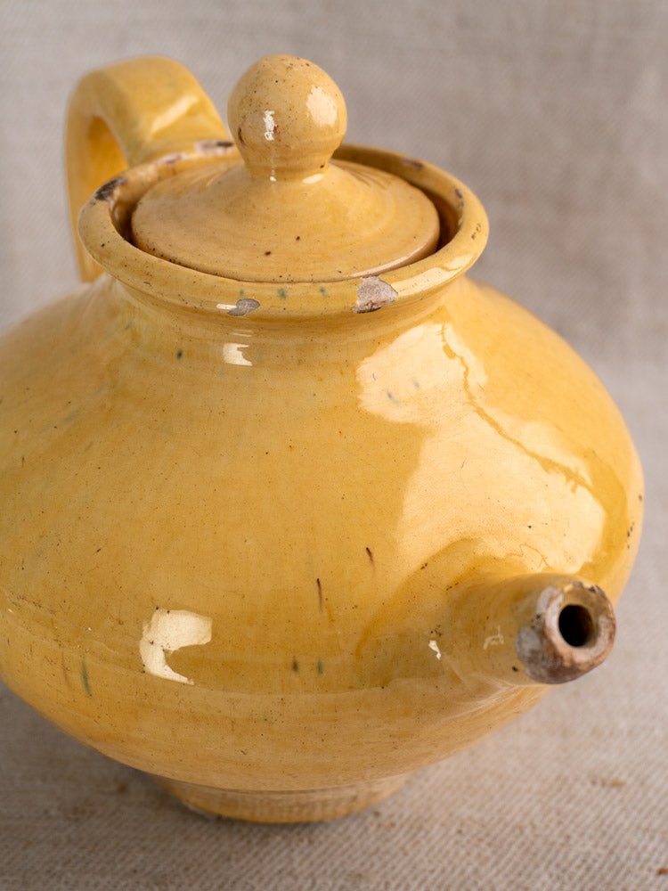 Antique French yellow glazed earthenware teapot with round lid, chipped rim, on textured beige fabric.