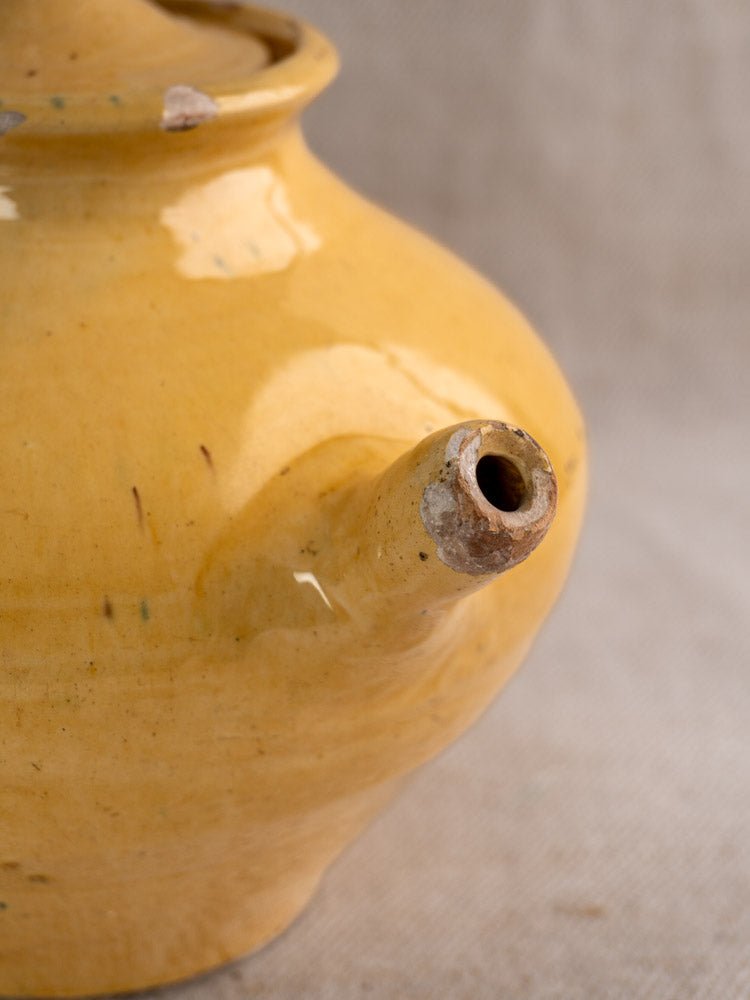 Antique French yellow glazed earthenware teapot with spout chips, neutral textured background.