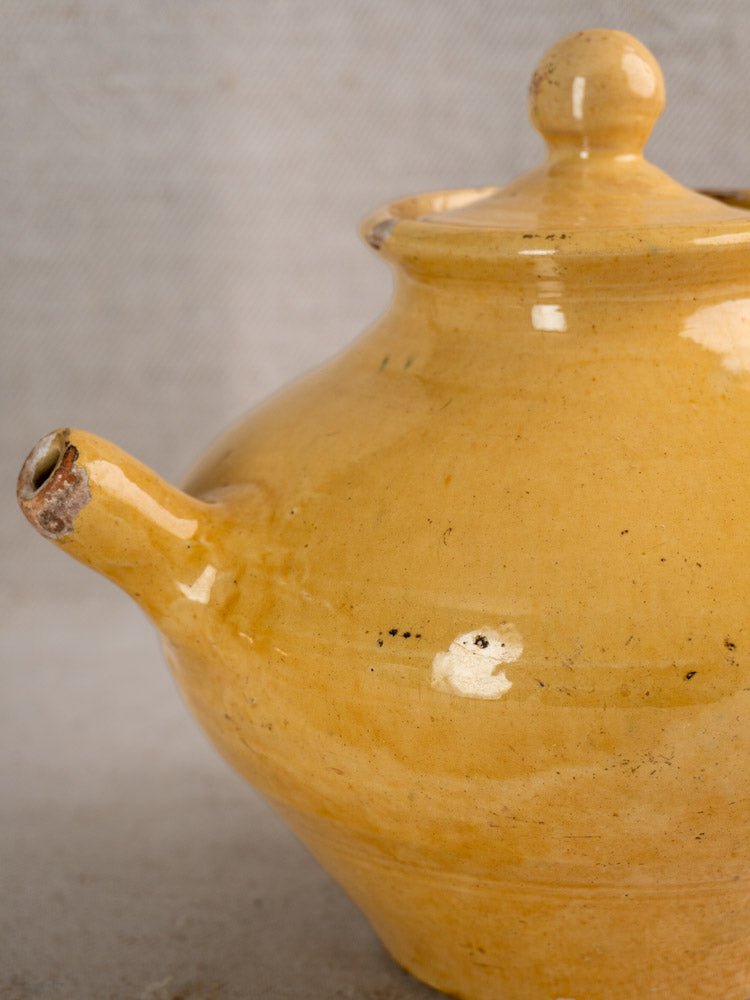 Antique French yellow glazed teapot with round lid and short spout on a neutral textured background.