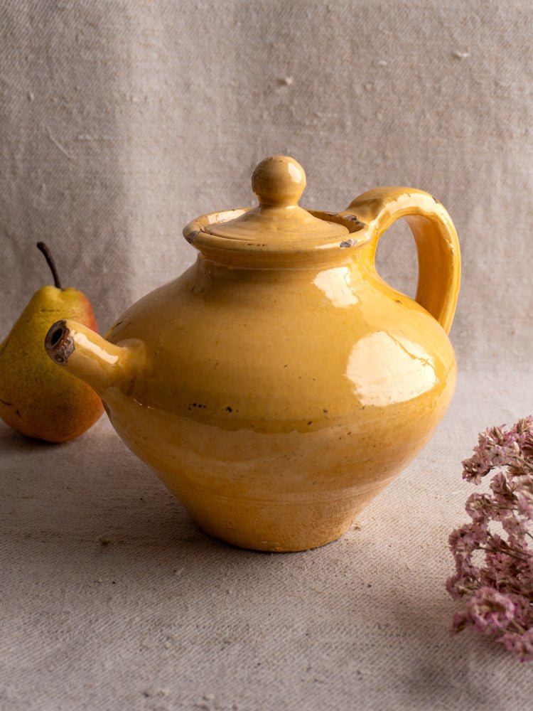 Antique French yellow glazed earthenware teapot with round handle and lid on beige cloth, pear, pink flowers nearby.