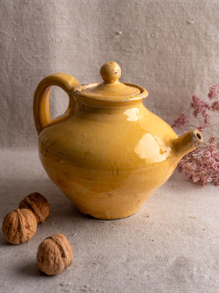 Antique French yellow glazed earthenware teapot on textured surface with walnuts and dried pink flowers.