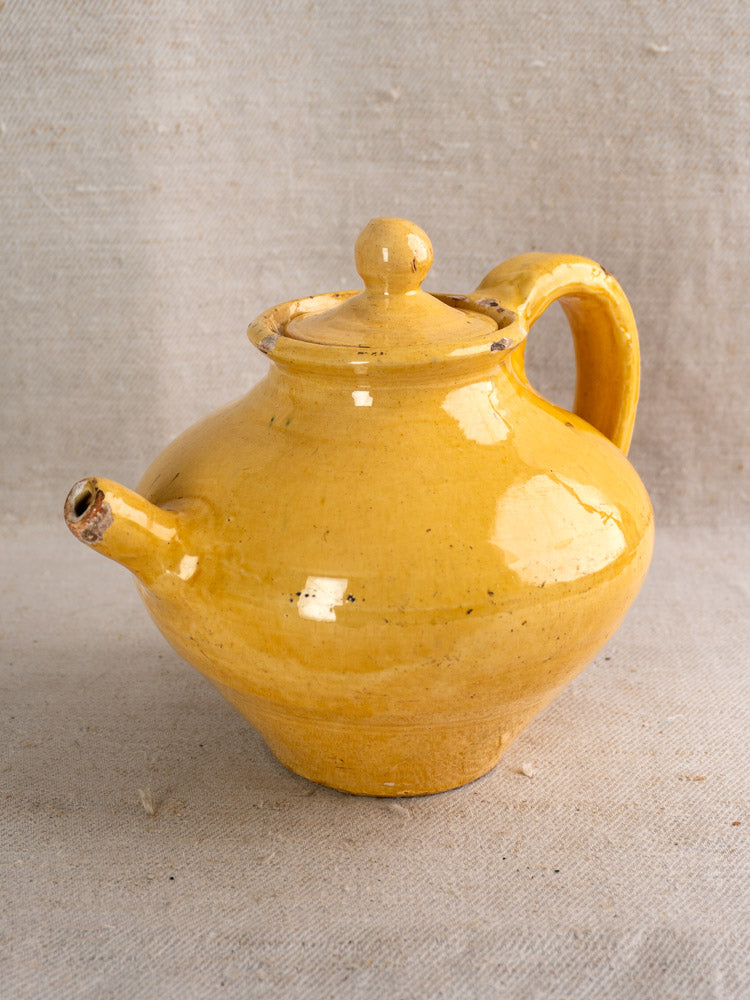 Antique French yellow glazed teapot with round lid and curved handle on beige fabric background.