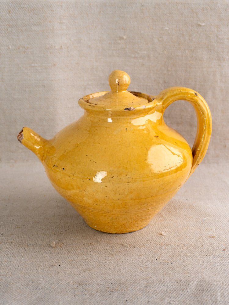 Antique French yellow glazed earthenware teapot with rounded body and curved handle on beige fabric.
