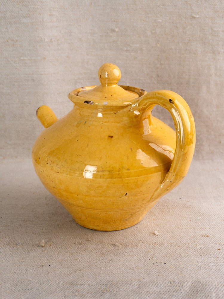Antique French yellow glazed teapot with curved handle and lid on textured beige fabric background.