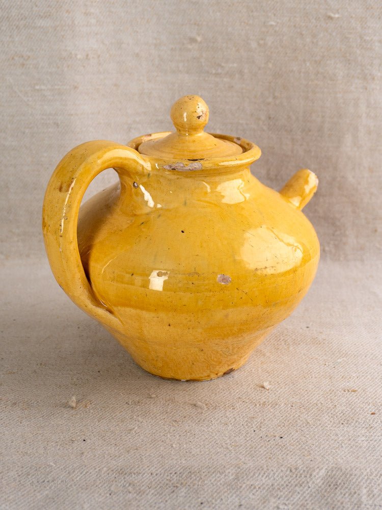 Antique French yellow glazed earthenware teapot with round lid and curved handle on beige fabric.