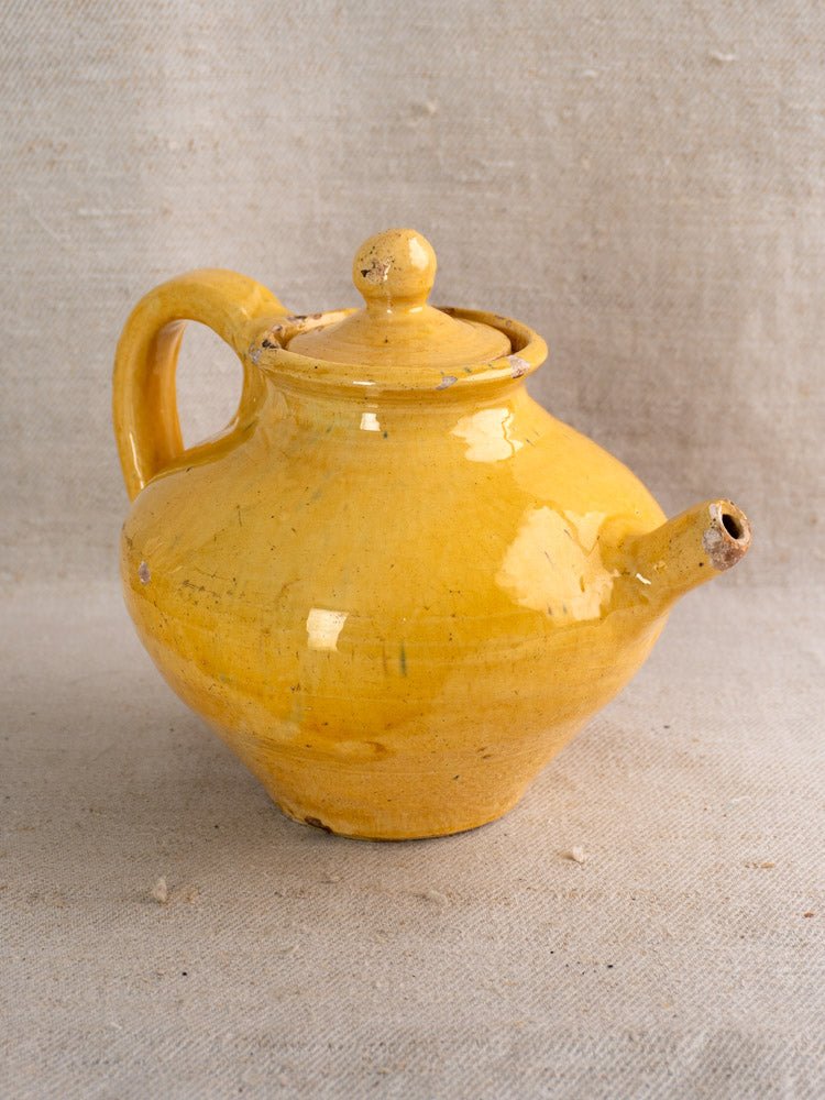 Antique French yellow glazed earthenware teapot with round lid and curved handle on beige fabric.