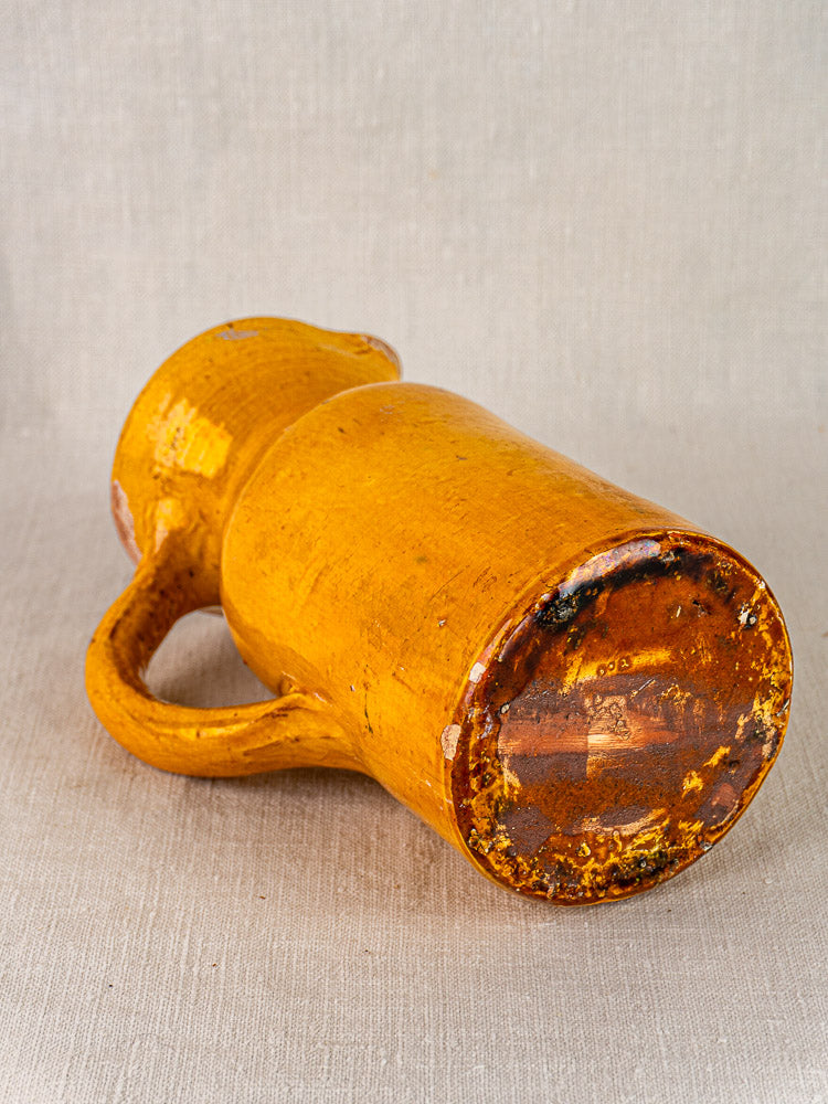 Antique French yellow-glazed terracotta pitcher with rounded base and curved handle on beige fabric.