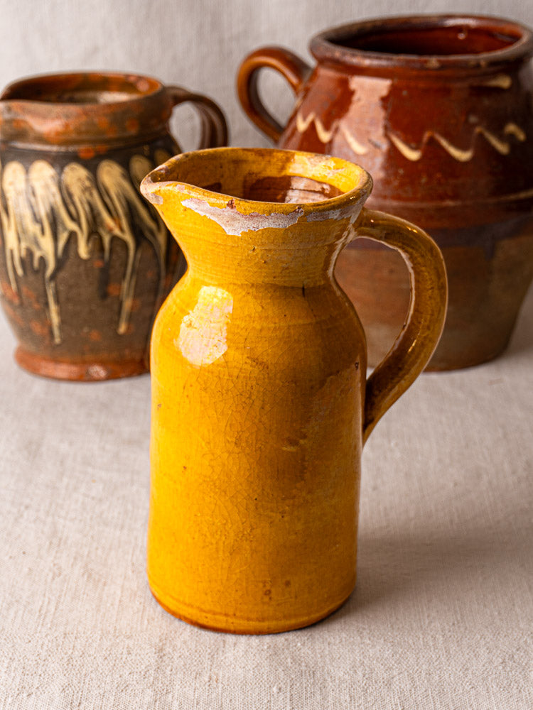 Antique French Pichet Jaune, yellow glazed terracotta pitcher with chipped edges, before two brown pottery jugs on textured fabric.
