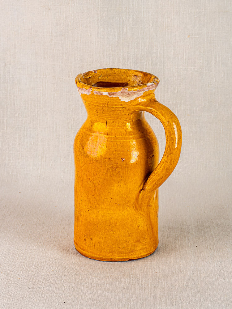 Antique French yellow glazed terracotta jug with handle and chipped rim on neutral fabric.