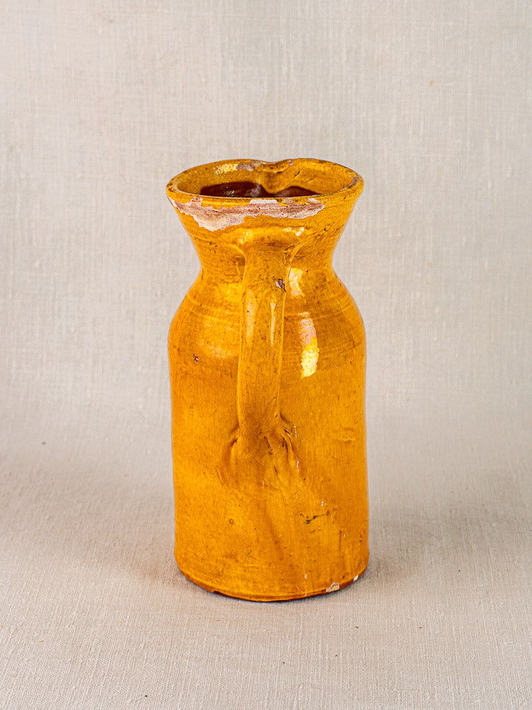 Antique French Pichet Jaune, tall yellow-glazed terracotta pitcher with handle and narrow neck on fabric background.