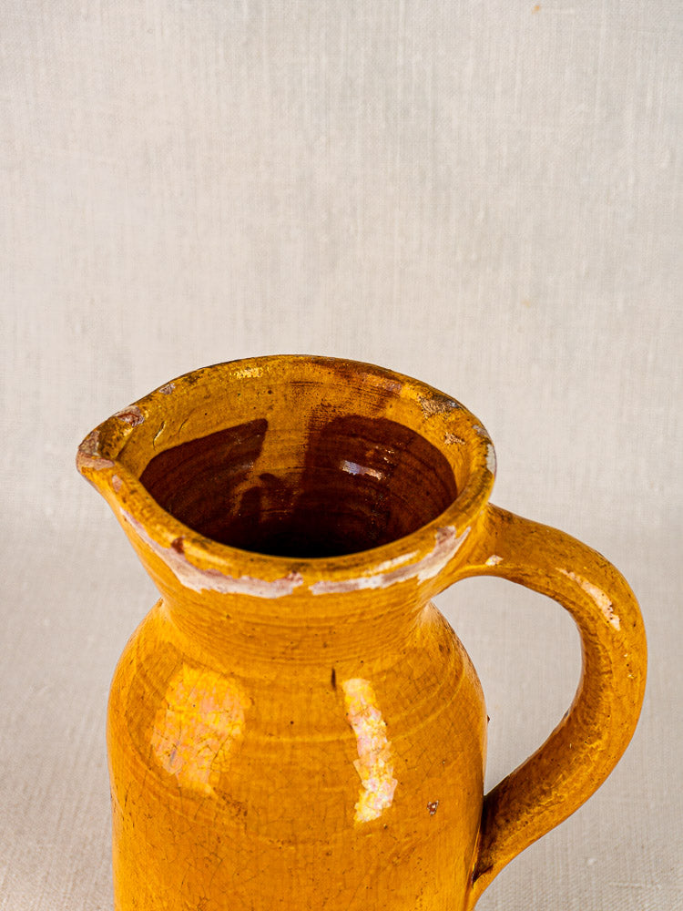 Antique French Pichet Jaune, 19th-century yellow glazed terracotta pitcher with handle and spout on neutral fabric.
