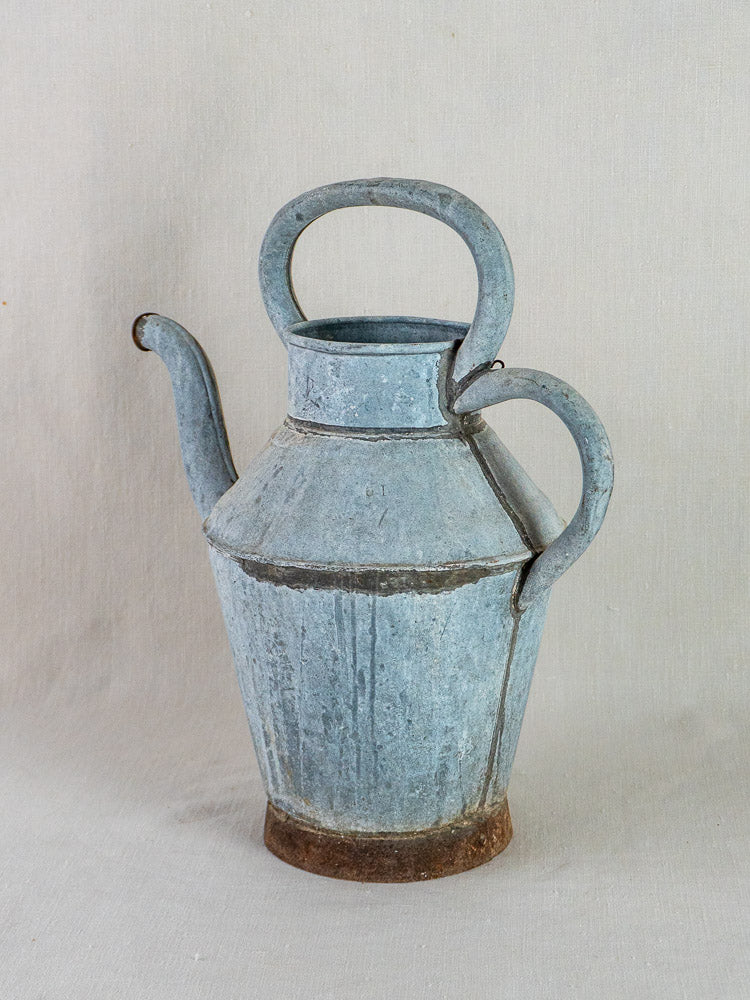 Antique French zinc broc à eau water pitcher with blue-gray patina, curved spout, and dual handles on beige background.