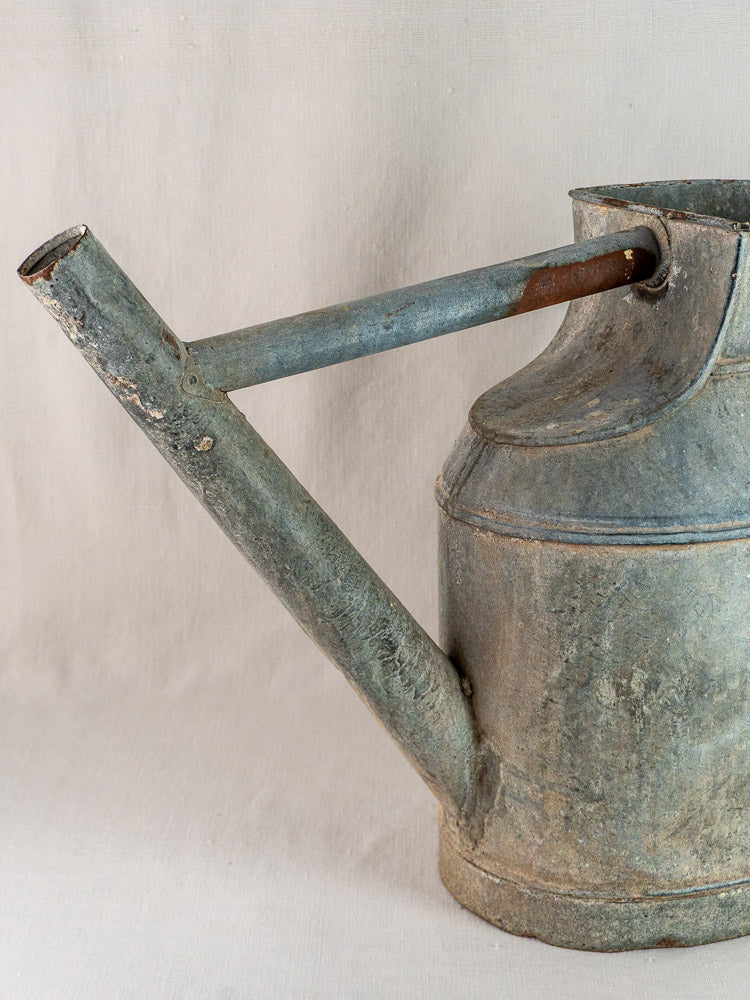 Large antique French zinc watering can with a long spout, weathered metal finish, plain beige background.