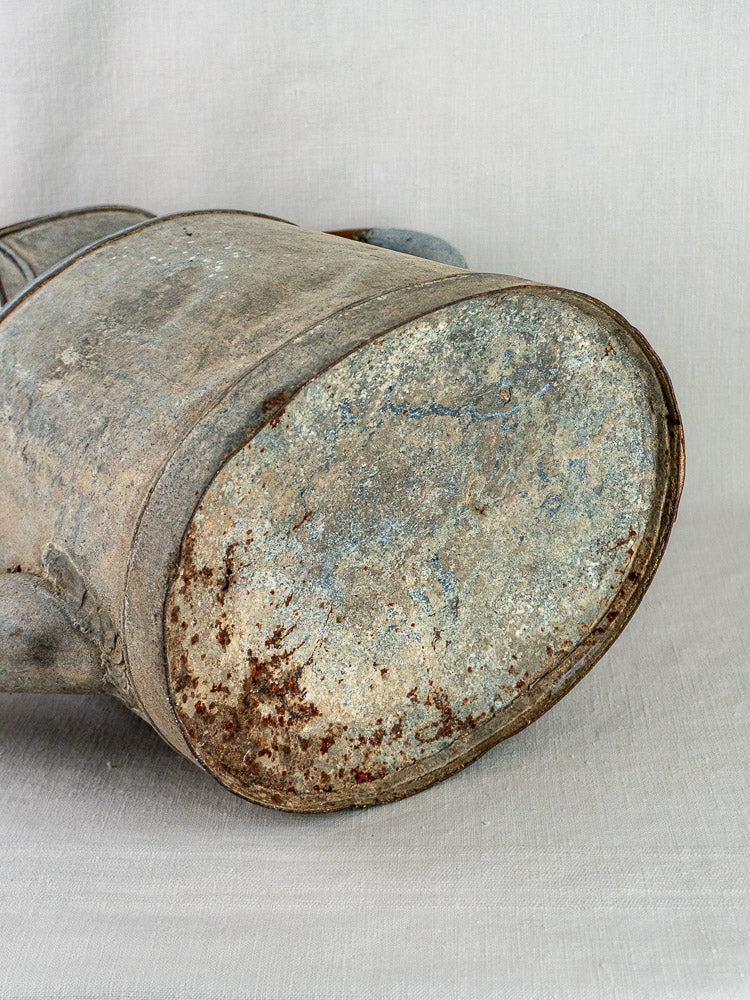 Close-up of the rusted base of a large French zinc antique market garden watering can on light fabric.