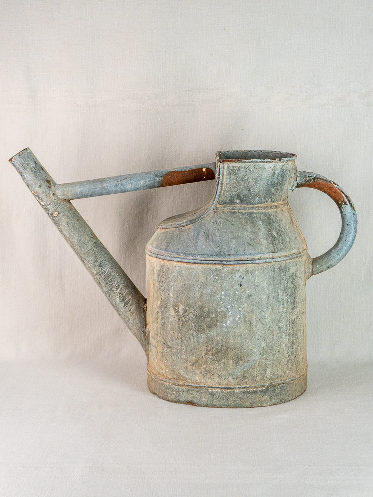 Large antique French zinc arrosoir with elegant handle and long spout on a beige backdrop.
