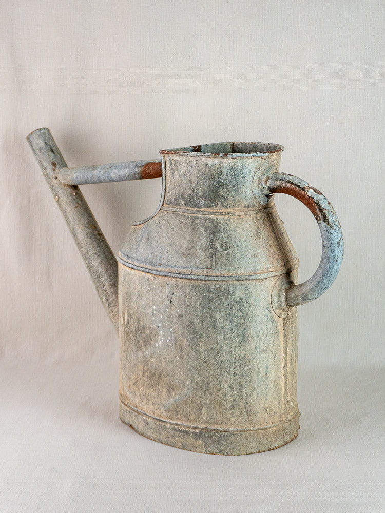 Large antique French zinc watering can with side handle and long spout on neutral fabric backdrop.