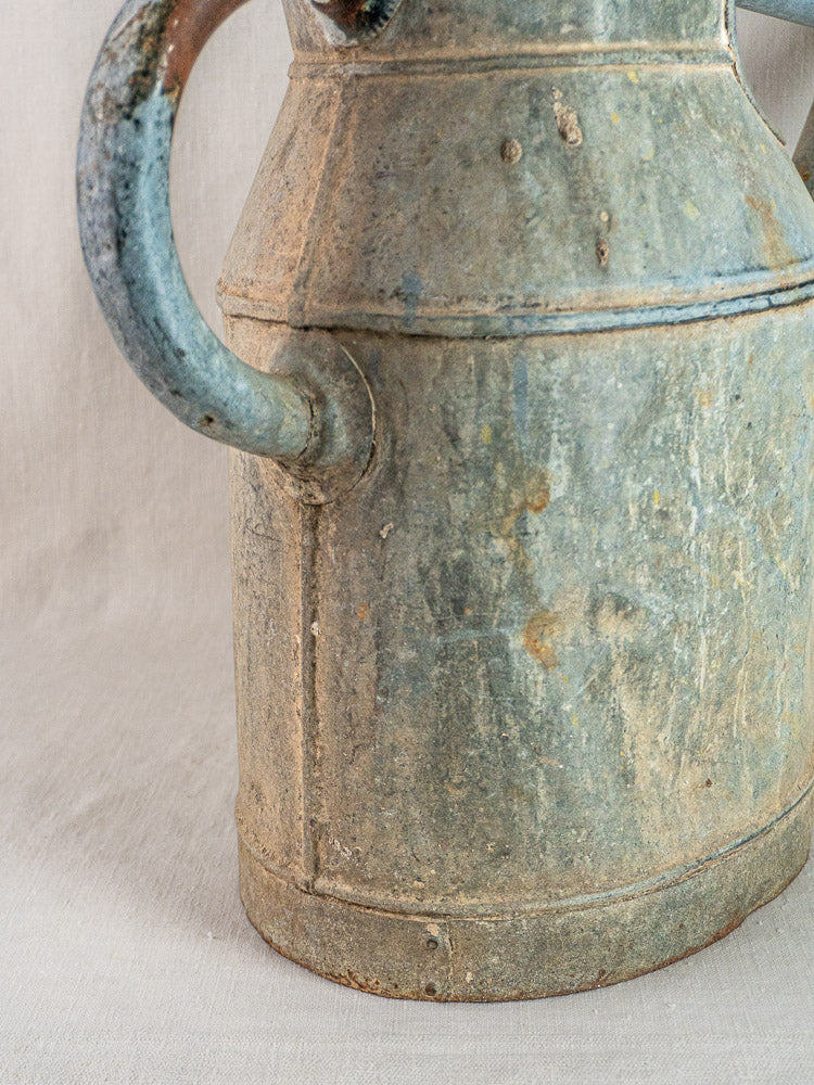 Large antique French zinc watering can with curved handle, aged patina, on neutral fabric background.