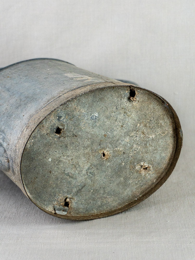 French zinc watering can planter with aged cylindrical base and drainage holes on neutral fabric.