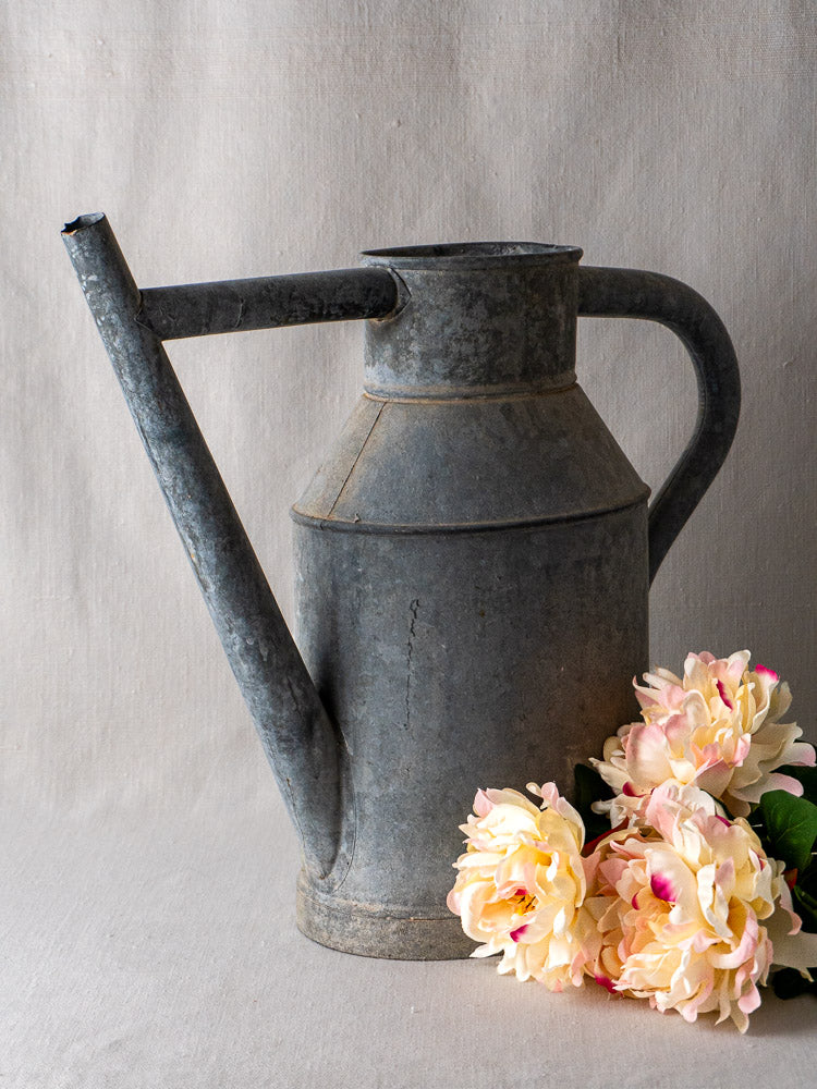 Antique French zinc long-spout watering can planter with light pink artificial flowers at base.
